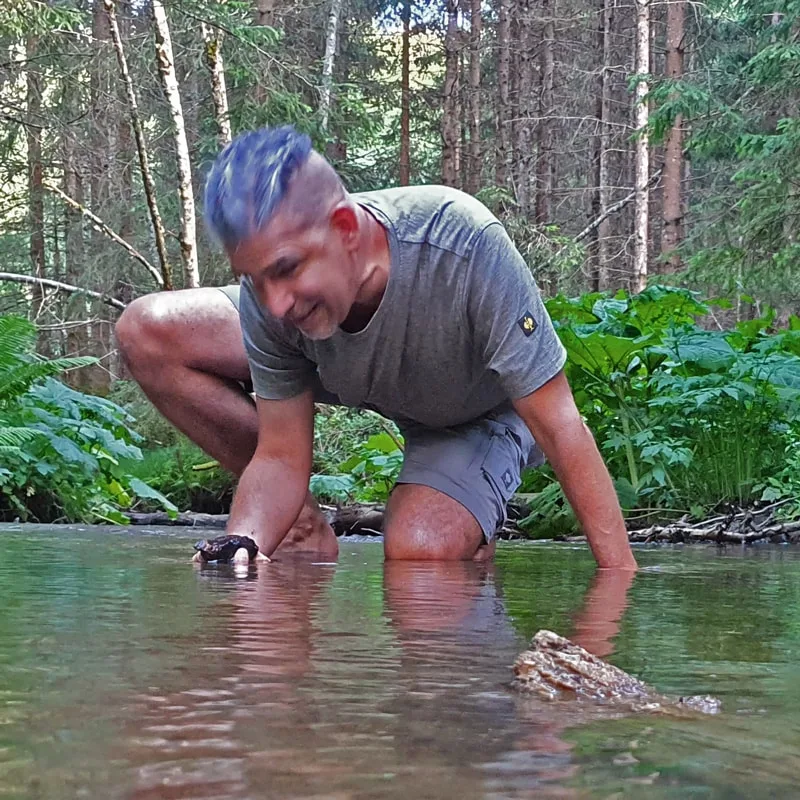 Matthias kniet barfuß in einem klaren Waldbach, beugt sich mit einem Lächeln nach vorne und berührt achtsam das Wasser – umge