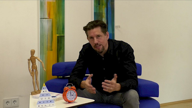Mann im schwarzen Hemd sitzt auf blauem Sofa und gestikuliert beim Sprechen