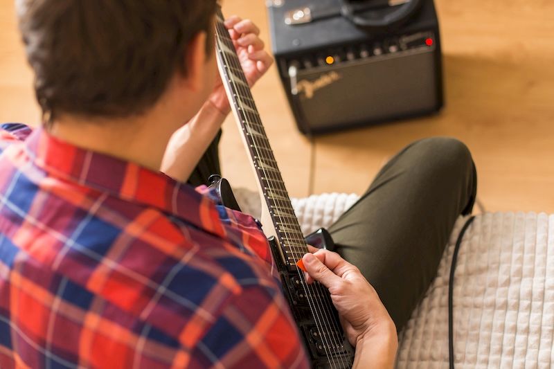 Schnellstart Rockgitarre – Die Grundlagen in 6 Monaten 2 Mann von hinten spielt E-Gitarre, rot-blau kariertes Hemd, Gitarrenverstärker im Hintergrund