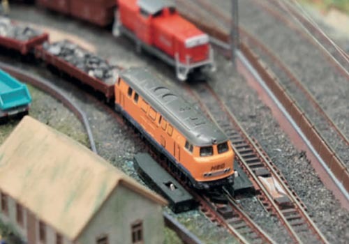 Modellbahnanlage mit orangeroter Lokomotive auf Gleisen, Häuser im Hintergrund