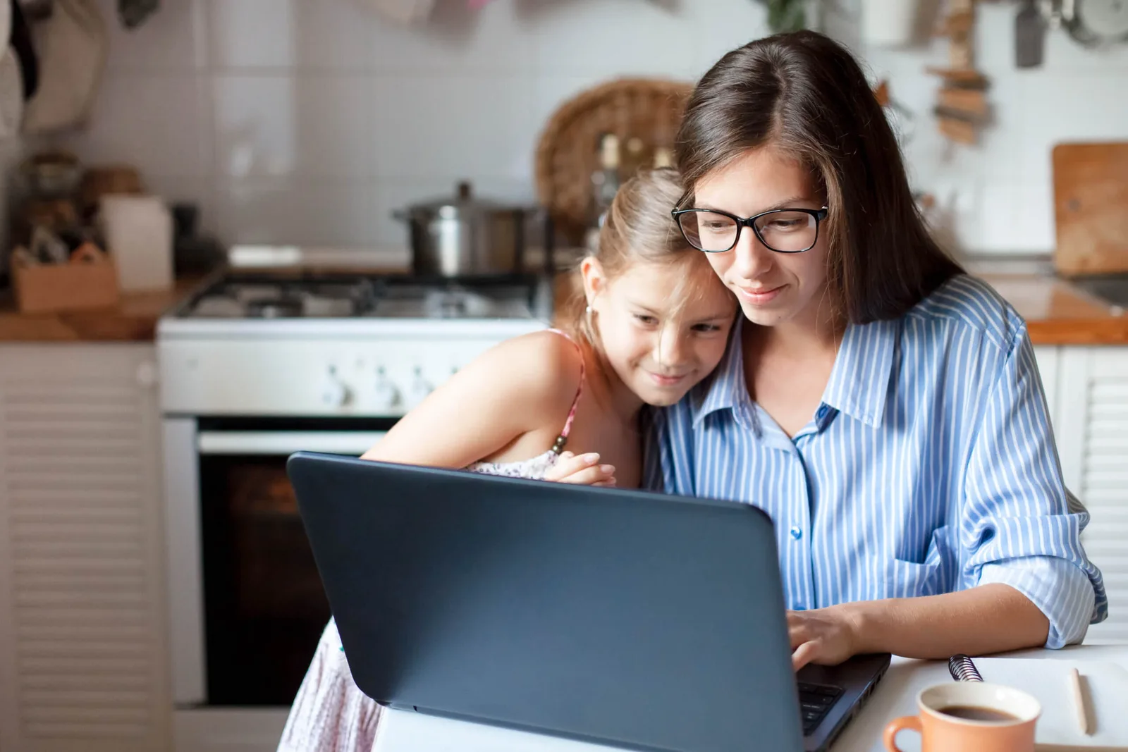 Frau mit Brille und Kind am Laptop in heimeliger Küche, gemeinsam lernend