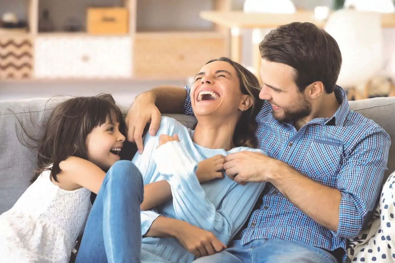Familie mit zwei Kindern und Eltern sitzt lachend auf grauem Sofa