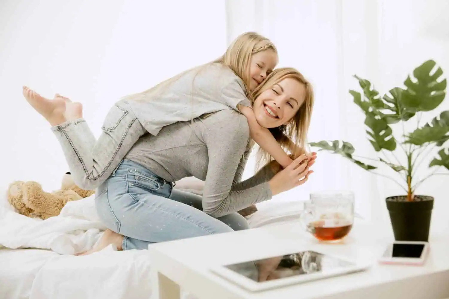 Mutter mit Tochter auf dem Arm im sonnigen Zimmer, mit Grünpflanze und Tasse