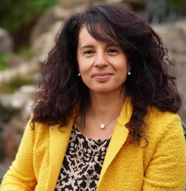Porträtfoto von Dorina Insalaco mit dunklen gewellten Haaren, gelbem Blazer und gemustertem Top vor unscharfem Steinhintergru