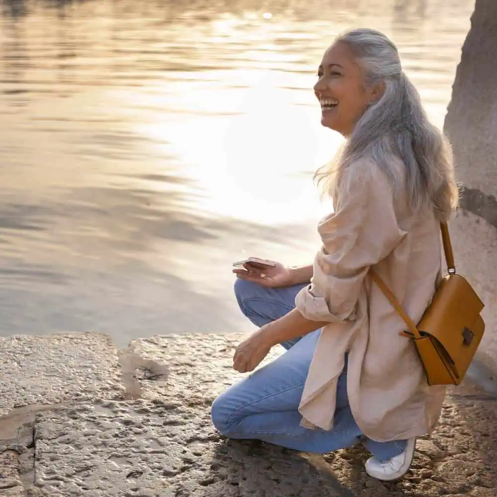 Frau mit grauem Haar sitzt lächelnd am Wasser, trägt beige Blazer und braune Umhängetasche