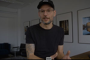 Videostandbild: Mann mit dunklem Shirt und Cap spricht in die Kamera im hellen Raum