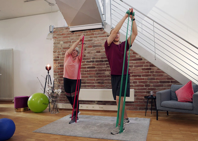 Trainerin und ältere Frau mit Fitnessbändern bei Knietraining, Loftwohnung