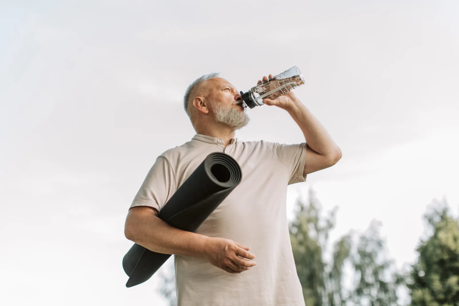 Älterer Mann mit Yogamatte und Wasserflasche beim Outdoor-Training vor blauem Himmel