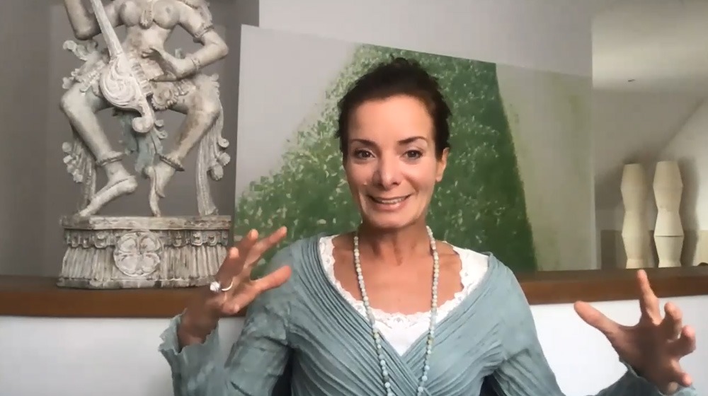Frau mit dunklem Haar in grauem Oberteil, gestikulierend vor modernem Hintergrund