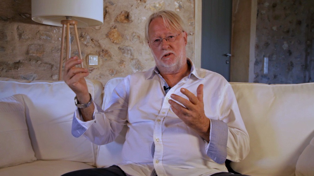 Dieter Broers auf Sofa mit gespreizten Händen, weißes Hemd, sprechend