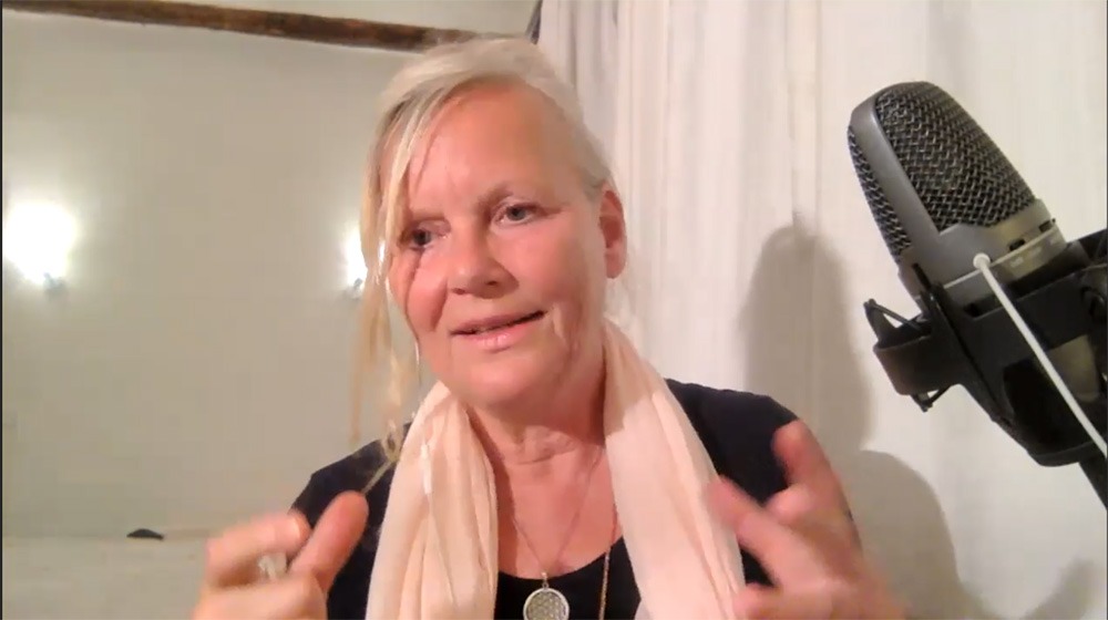 Sabine mit Mikrofon, rosa Tuch um Schultern, gestikulierend