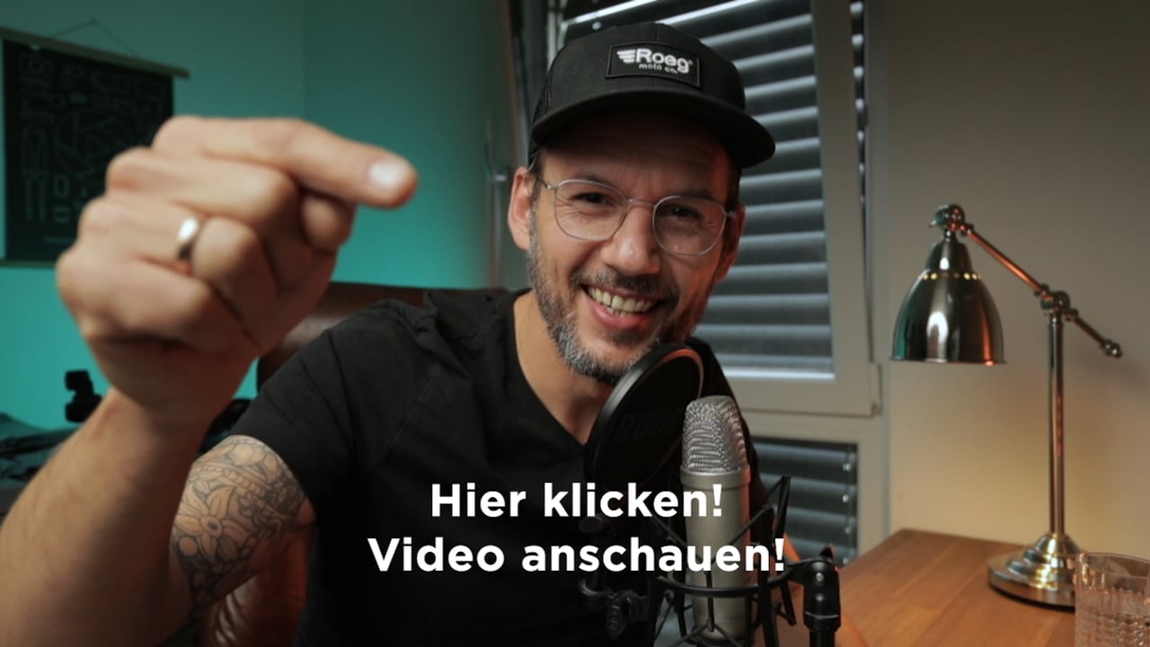 Mann mit Kappe und Brille am Mikrofon zeigt auf Kamera mit Text 'Hier klicken! Video anschauen!'