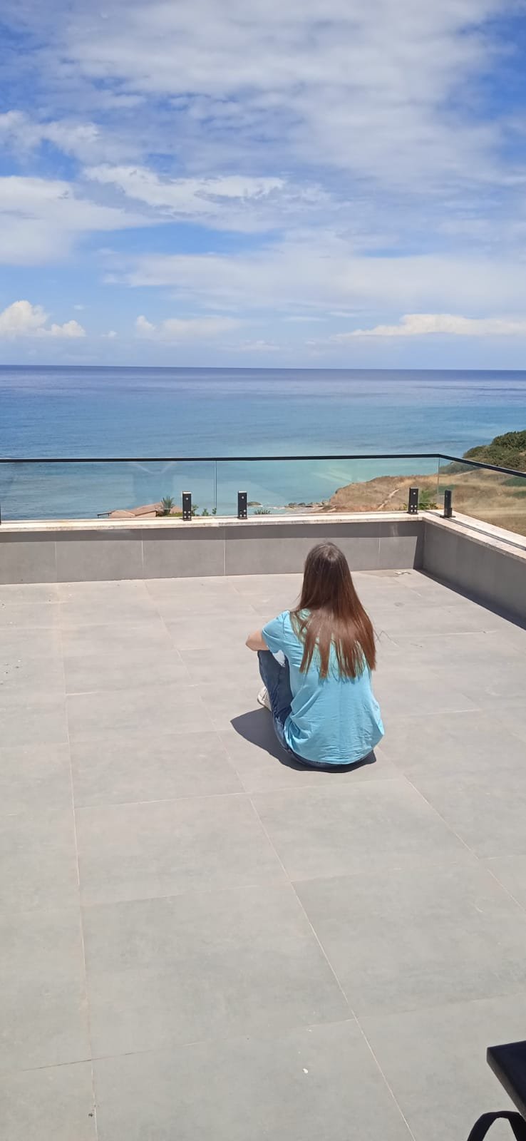 Frau in blauem Shirt sitzt auf Dachterrasse mit Meerblick und klarem Himmel