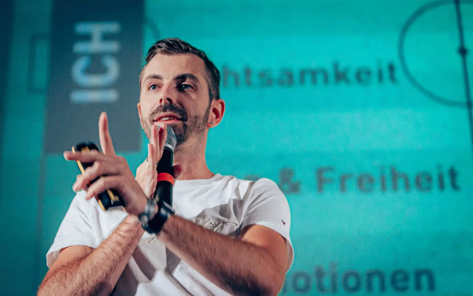 Oliver Albrecht spricht in weißem Shirt mit Mikrofon auf türkisfarbenem Hintergrund Keynote
