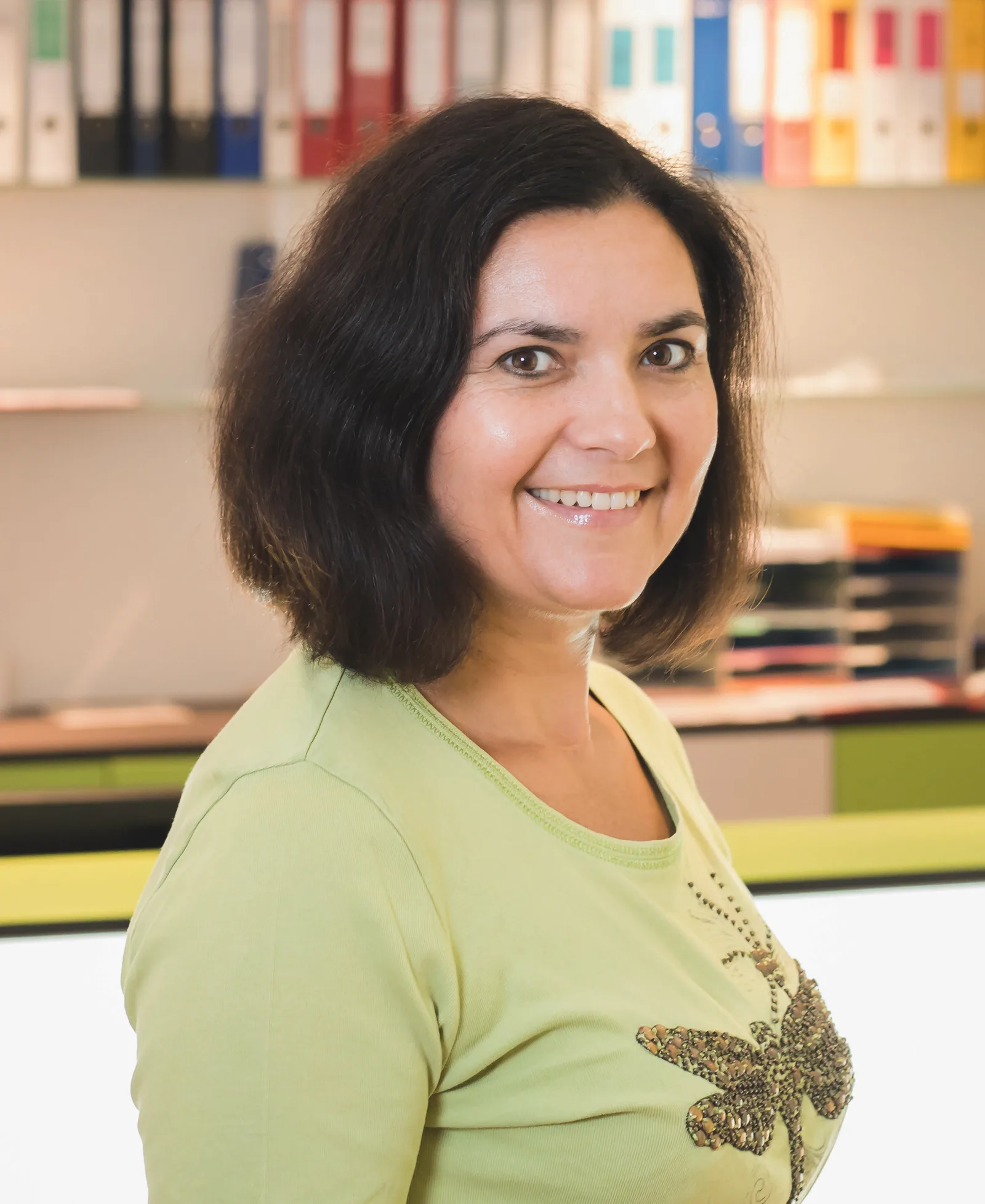Porträt einer Frau mit schwarzem Haar im grünen Shirt, lächelnd vor buntem Bücherregal