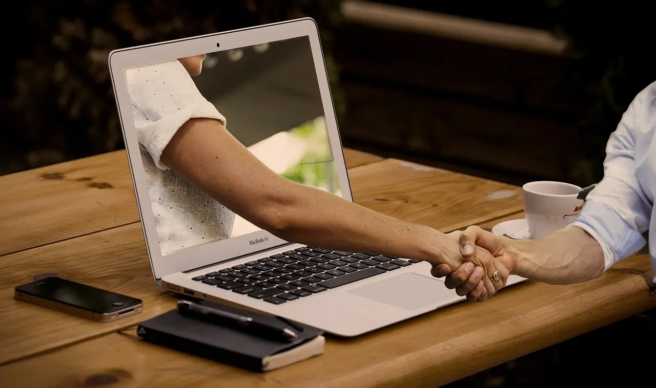 Person beim virtuellen Handshake über Laptop-Bildschirm am Holztisch