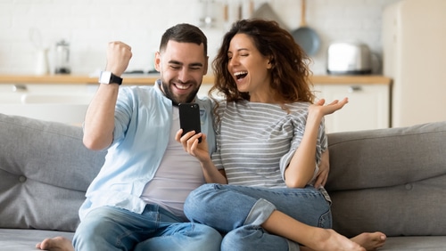Junges Paar sitzt auf Sofa und jubelt über Smartphone-Erfolg