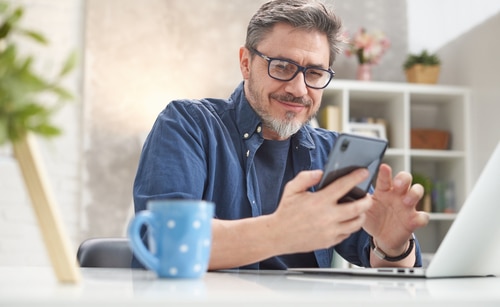 älterer Mann mit Brille sitzt am Schreibtisch und prüft Smartphone-News