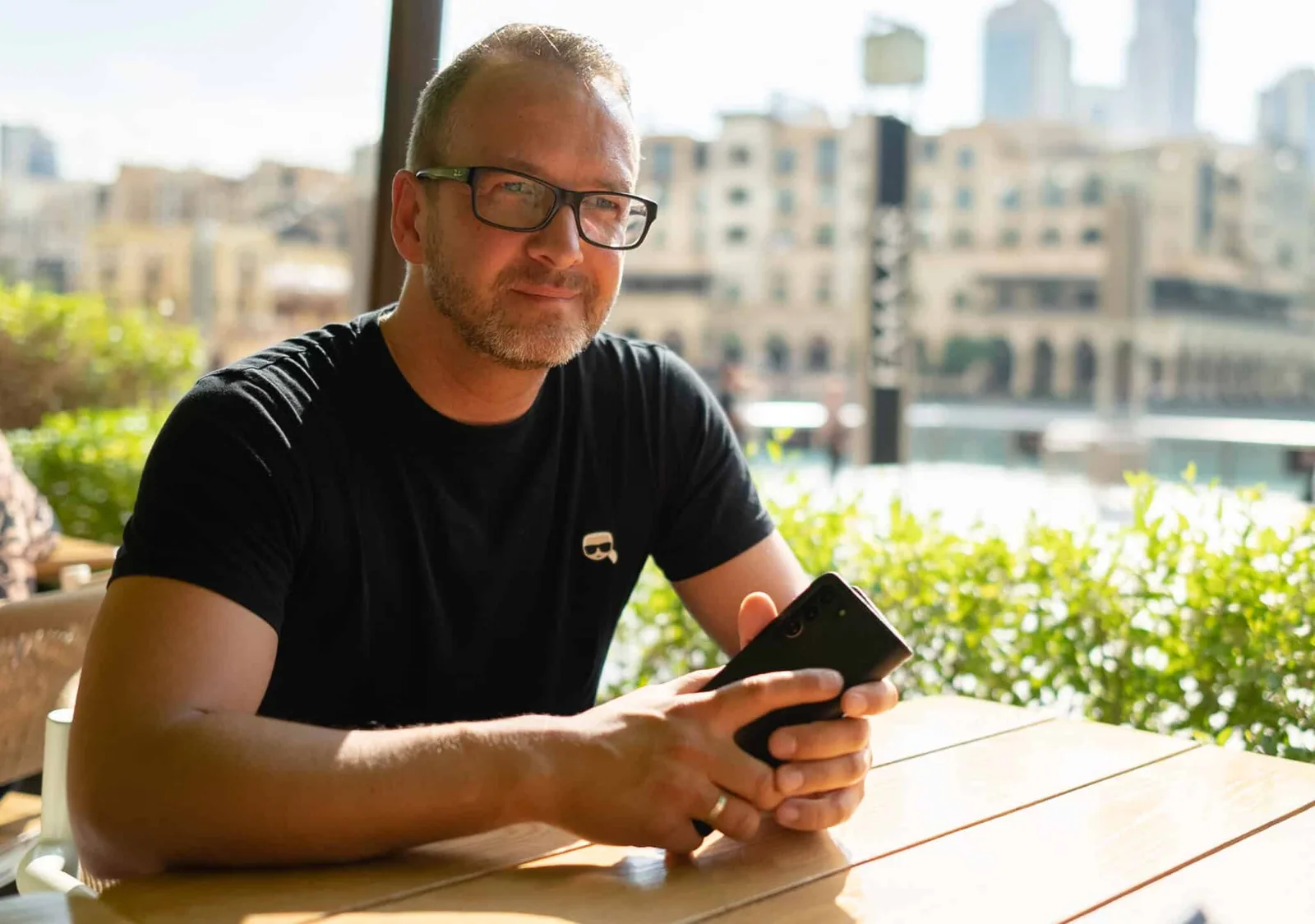 Mann mit Brille hält Smartphone auf Terrasse mit Stadtblick im Hintergrund, Smartphone Business Webinar 2
