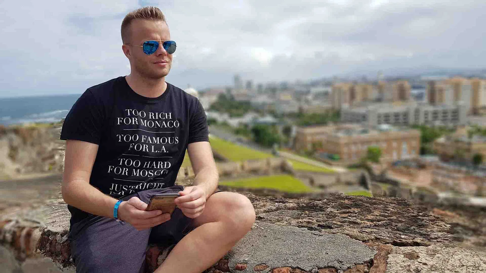 Gunnar Kessler sitzt auf einer Felskante mit Smartphone, trägt Sonnenbrille, Küstenlandschaft im Hintergrund
