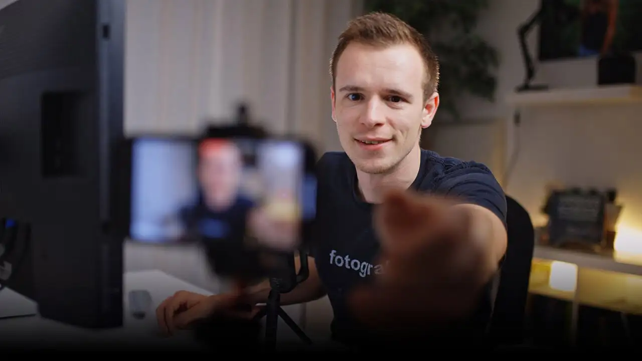 Junger Mann in Innenraum mit Smartphone, lächelt in Kamera, Filmset im Hintergrund