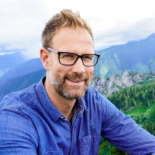 Porträt von Mann mit Brille im blauen Jeanshemd vor Berglandschaft-Hintergrund