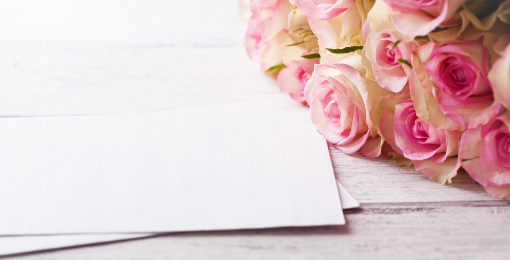 Weiße Grußkarte neben Strauß aus rosa und cremefarbenen Rosen auf weißem Holzuntergrund