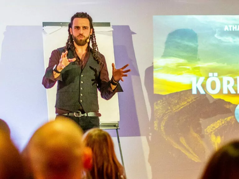 Mann mit langen Dreadlocks und Weste gestikuliert auf der Bühne vor Publikum