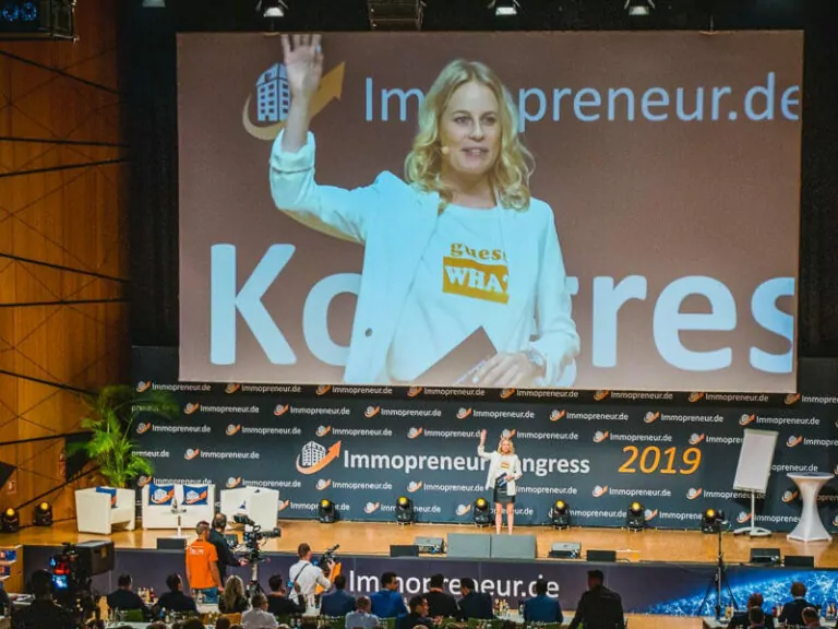 Melanie Siefert auf Bühne vor Leinwand mit Immopreneur.de Logo, trägt weißes Blazer