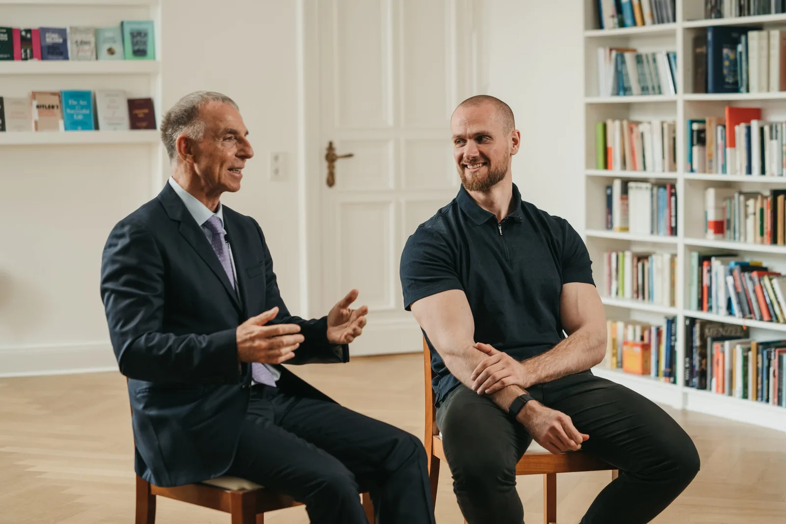 Zwei Männer im Gespräch auf Stühlen vor Bücherregal, einer im Blazer, einer in schwarzem Polo