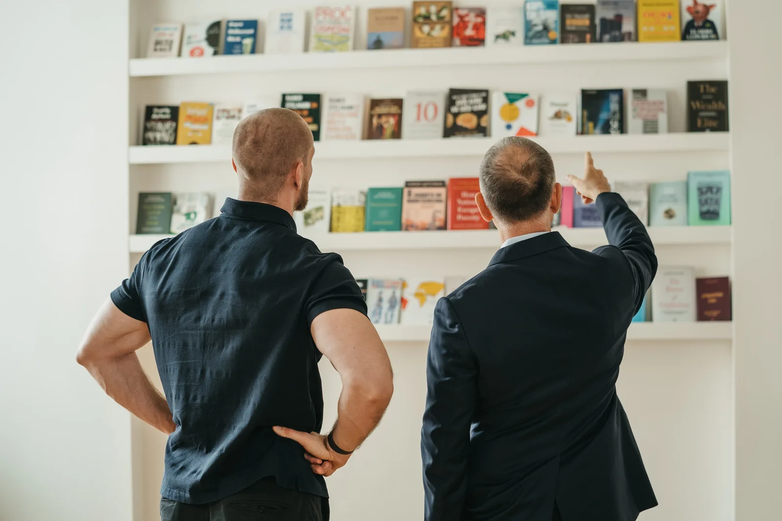 Zwei Männer von hinten vor Bücherregal, einer deutet auf Bücher