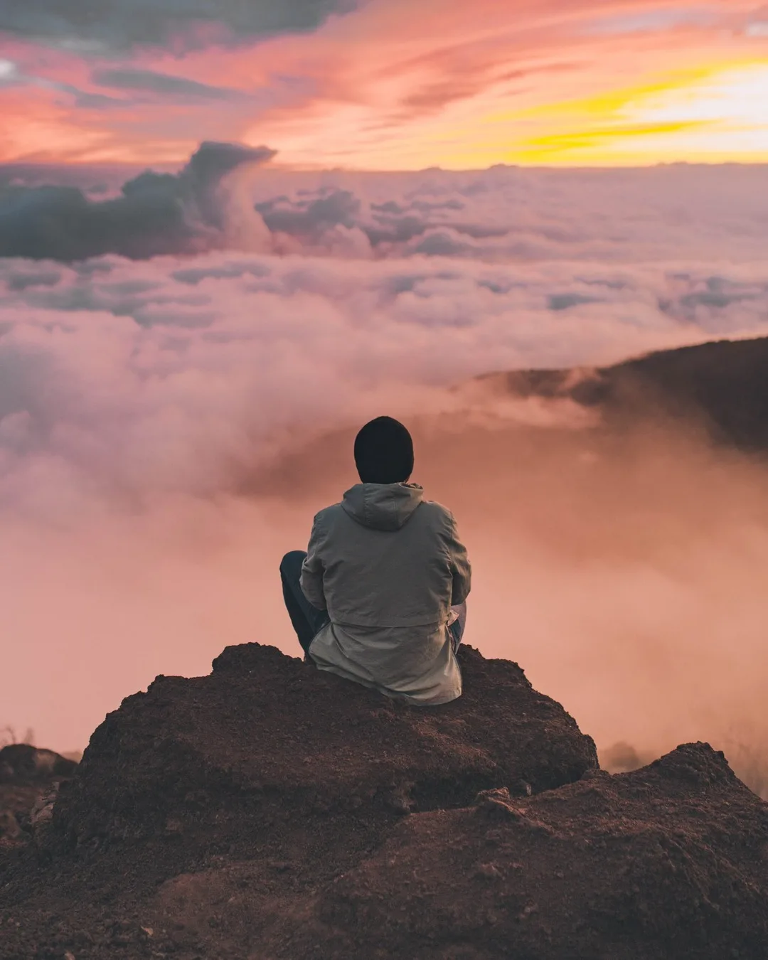 Person sitzt auf Felsen und schaut auf Wolkenmeer bei Sonnenaufgang