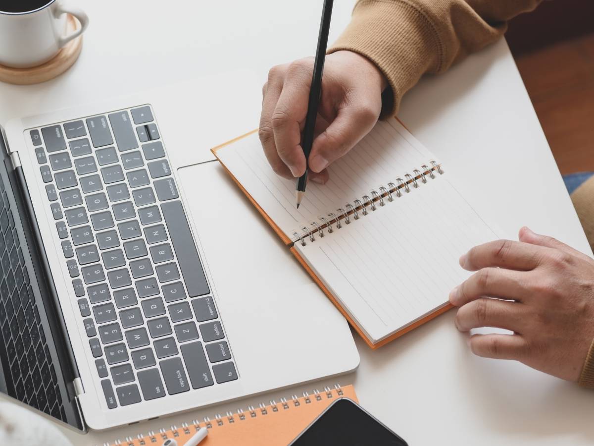 Person schreibt in Notizbuch neben Laptop, Kaffeetasse und Stift