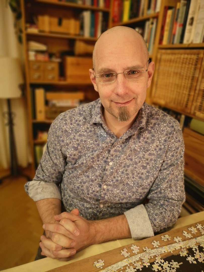 Porträt eines Mannes mit Brille vor Bücherregal, ein Puzzleteil in der Hand