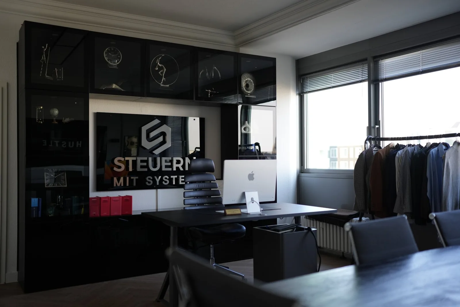Modernes Büro mit schwarzem Schrank, Steuergestaltung mit System Logo an Wand, Apple Monitor und schwarze Kleiderstange