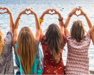 Vier Frauen von hinten am Strand bilden Herzform mit Armen