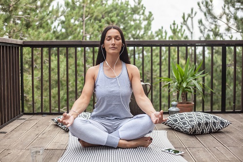 Frau in Yoga-Pose sitzt meditierend auf Holzterrasse mit Blick ins Grüne