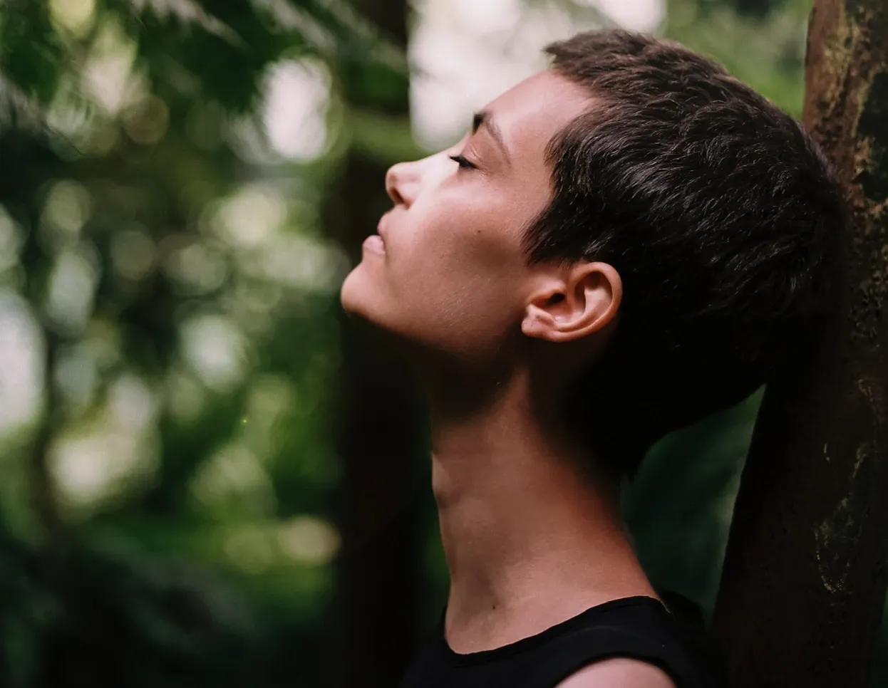 Stress Master – Videokurs für innere Ruhe von Maxim Mankevich 8 Junge Frau mit geschlossenen Augen lehnt an Baum im Wald, Frieden und Naturverbundenheit ausdrückend