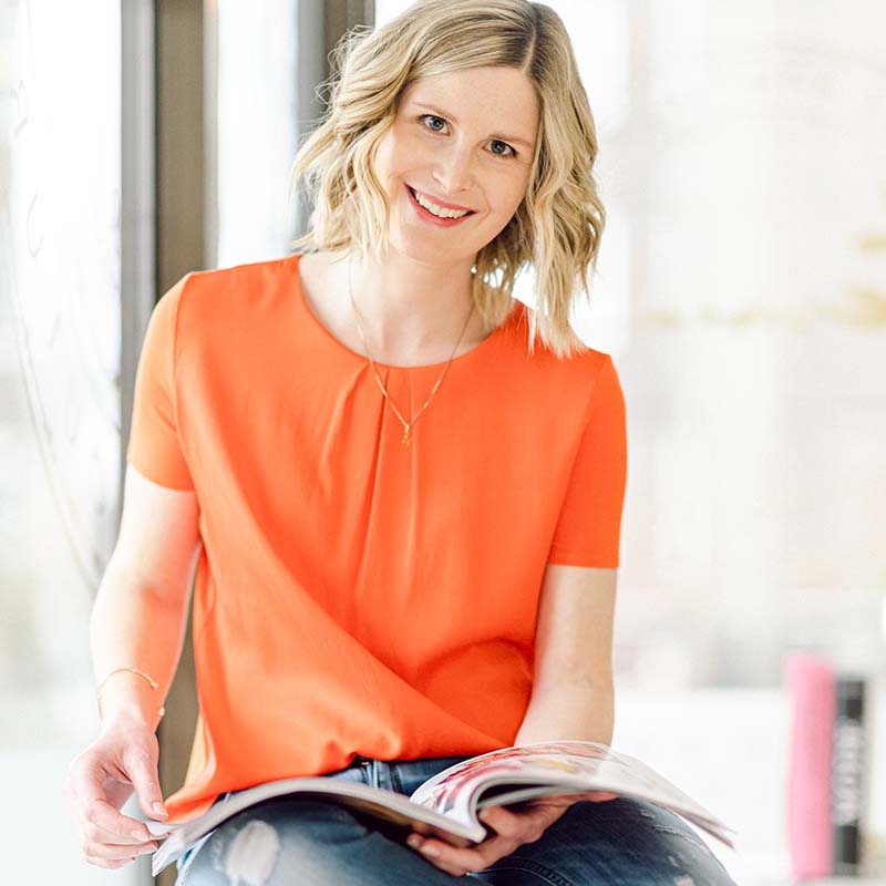 Portrait einer blonden Frau in orangefarbenem T-Shirt, hält aufgeschlagenes Magazin