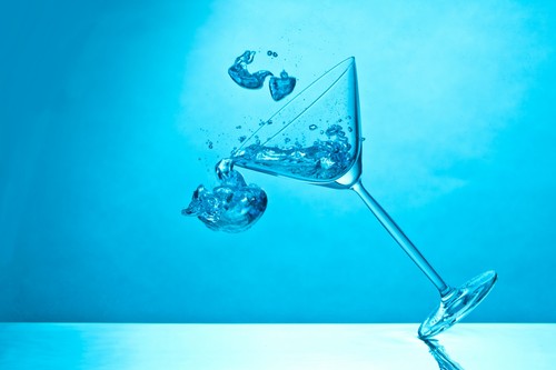 Umkippender Cocktailglas mit Eiswürfeln und Flüssigkeit auf blauem Hintergrund