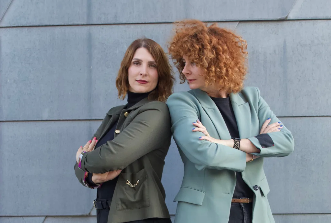 Zwei Frauen mit unterschiedlichen Haarfarben im professionellen Blazer vor grauer Wand