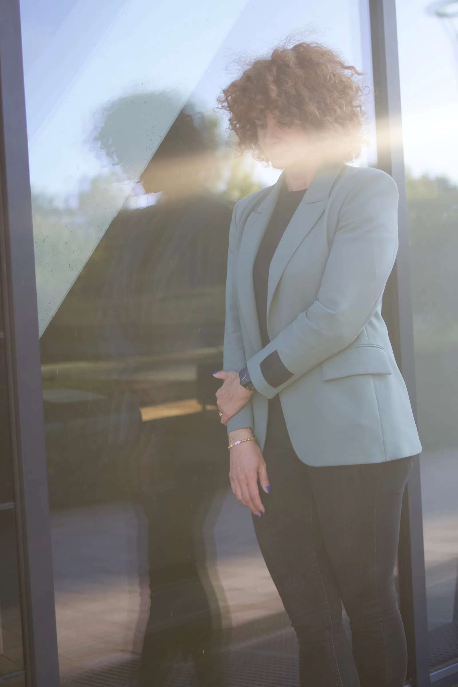 Frau im beigen Blazer blickt nachdenklich durch Fenster mit Spiegelung