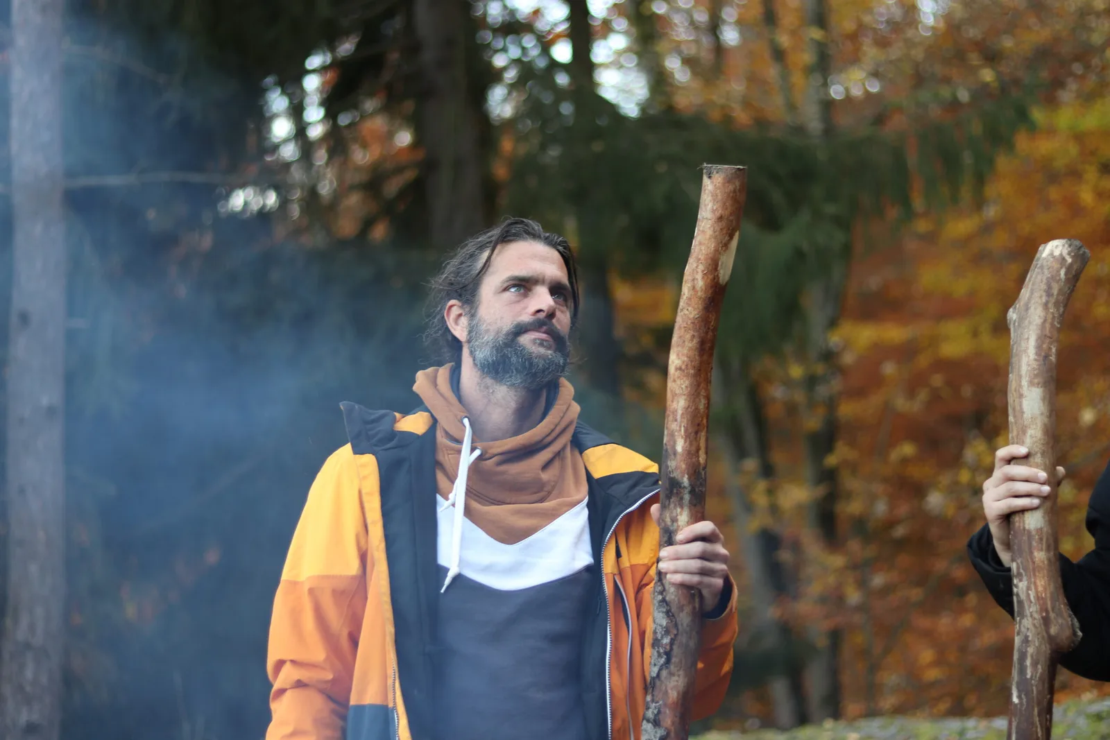 Mann mit braunem Schal und Jacke in Herbstwald, hält Ast, nachdenklicher Blick