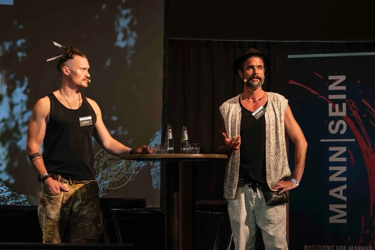 Zwei Männer auf Bühne, einer mit Dreadlocks in schwarzem Top, einer in Weste, neben Schriftzug Mannisein