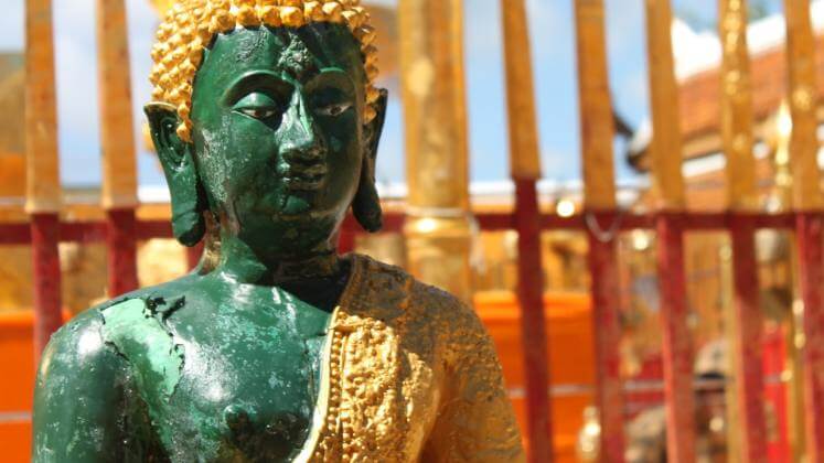 Grüne Buddha-Statue mit goldenem Kopfschmuck und rotem Tempel im Hintergrund, Thailand