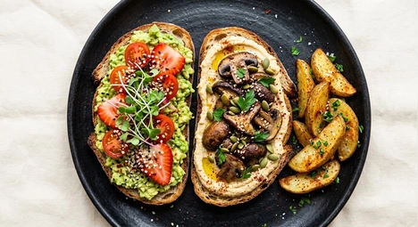 Zwei belegte Open-Toast-Varianten mit Avocado-Tomate und Pilzen sowie Kartoffelecken auf schwarzem Teller