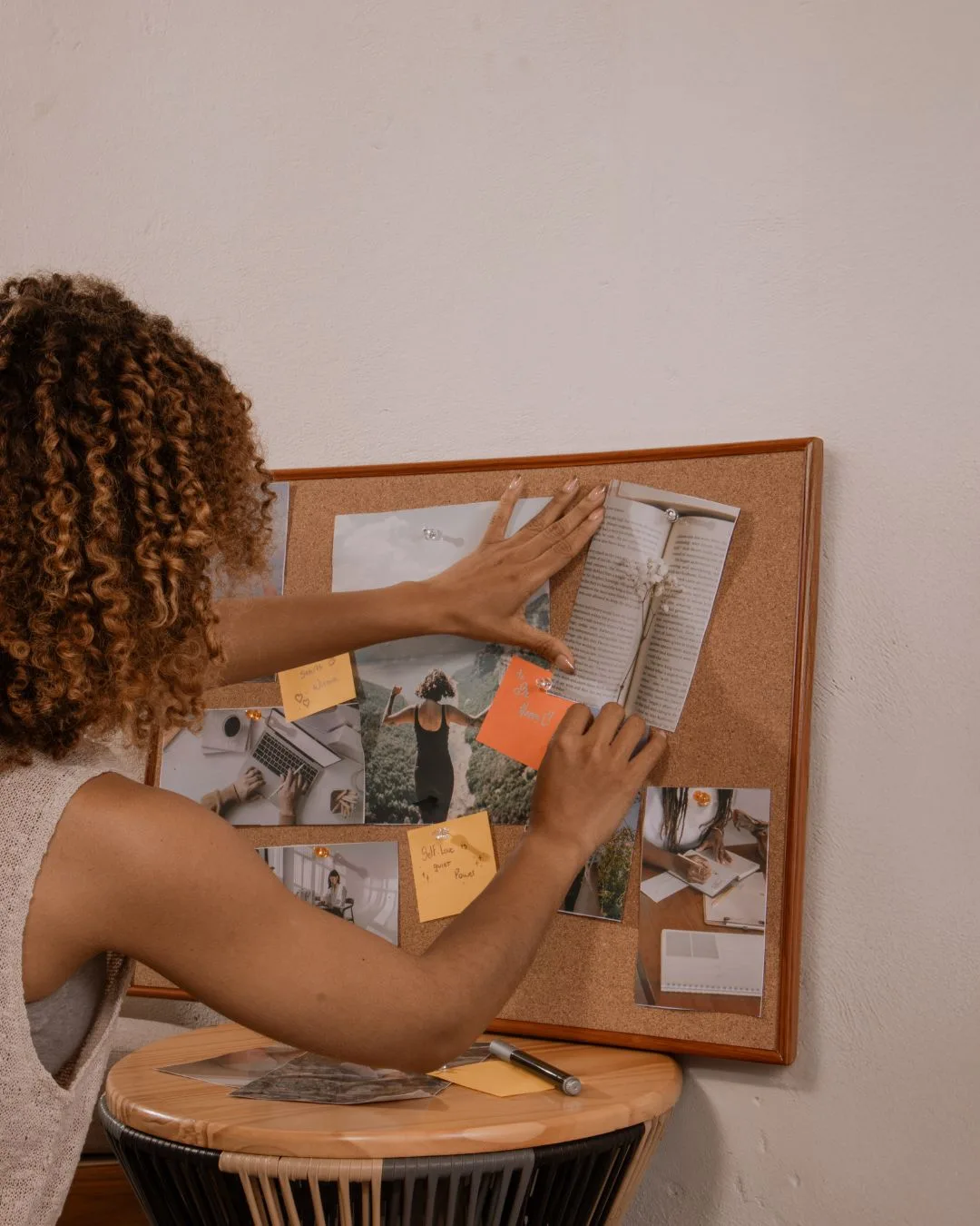 The Shine Vision Board System – Videokurs zur Manifestation 6 Frau von hinten ordnet Fotos und Notizen an einem Korkboard mit orangefarbenen Notizkarten