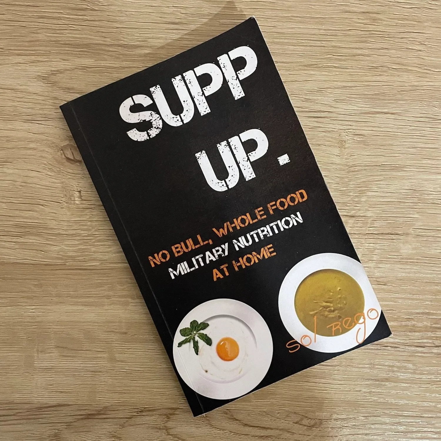 The SUPP UP. No Bull Nutrition At Home Field Guide 1 SUPP UP Taschenbuch-Cover auf Holztisch, schwarzes Design mit zwei Tellern mit Essen