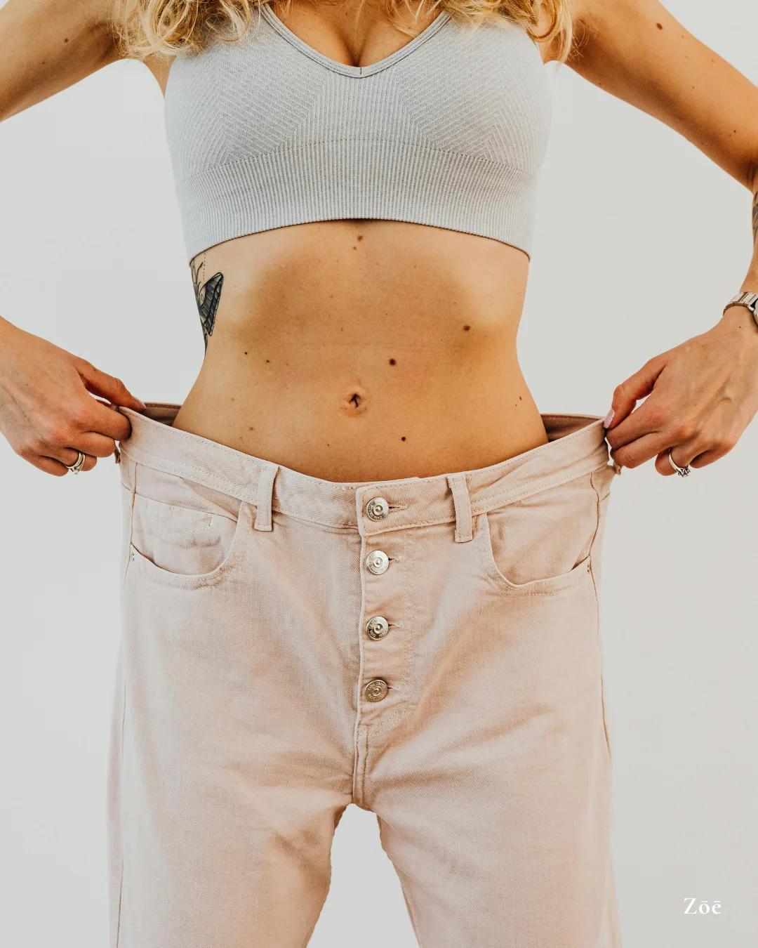 Frau zeigt flachen Bauch in beiger Jeans und hellgrauem Crop-Top, Fitness-Transformation Resultat