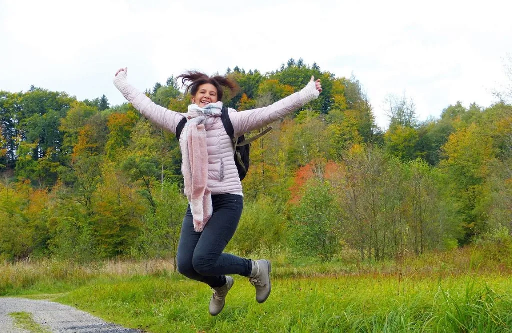 Frau springt jubelnd in die Luft mit ausgebreiteten Armen vor herbstlichem Wald